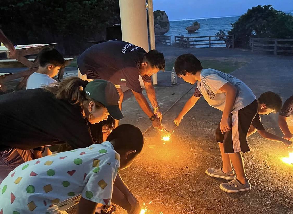 日が落ちた海辺で、スタッフと一緒に手持ち花火に火をつける子どもたち