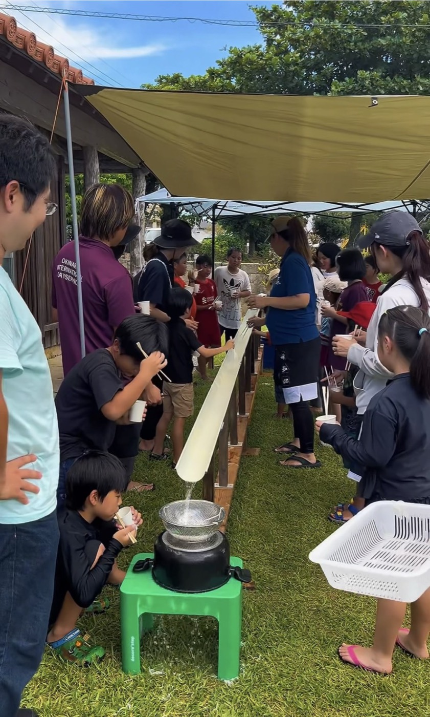 OKI HOUSEの庭に長い樋を設置し、多くの子どもと大人が流しそうめんを楽しむ様子