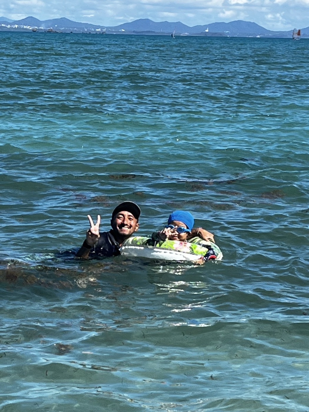 海の中で、浮き輪に乗った子どもを支えながらピースをするサポーター
