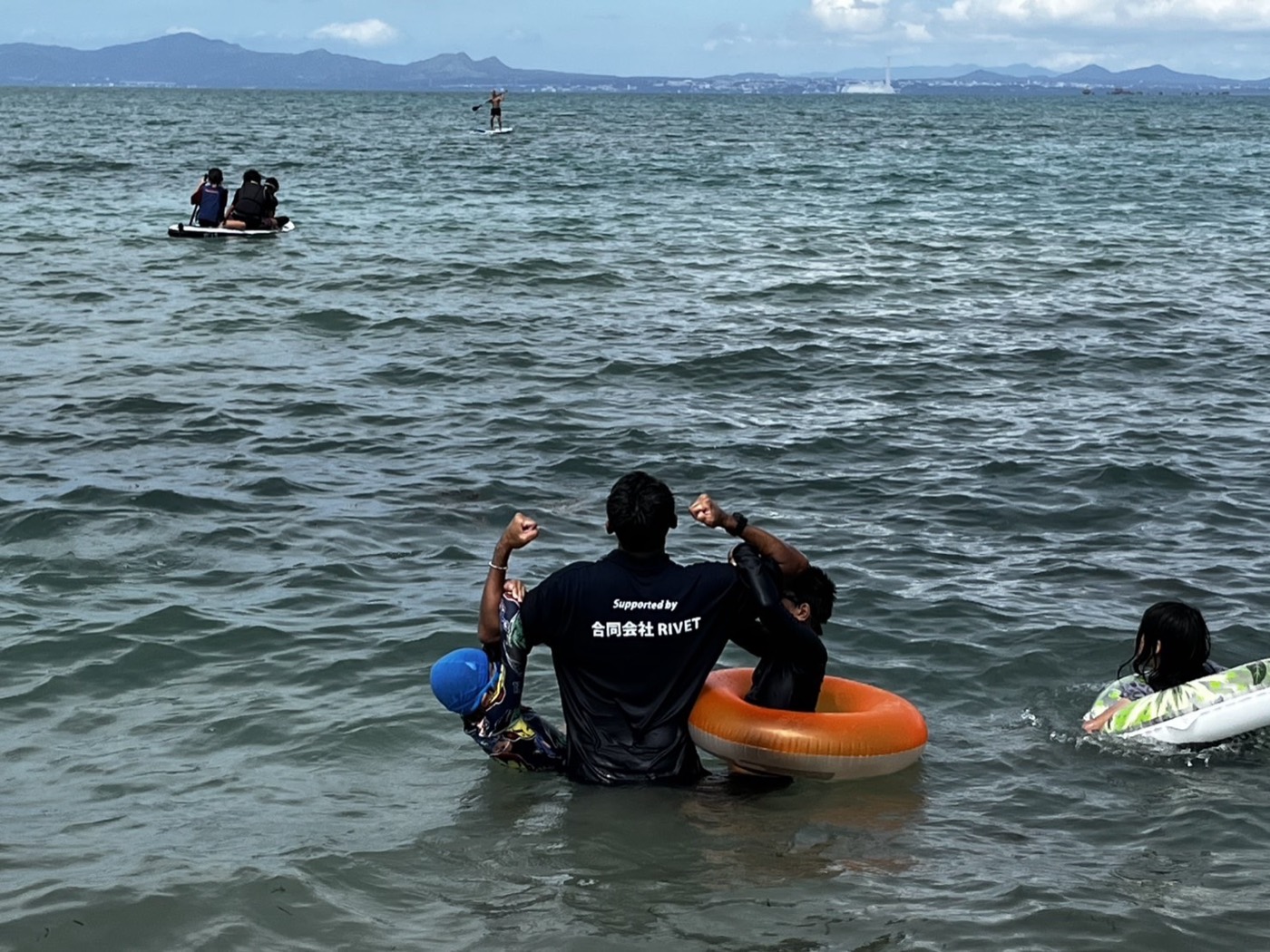 海の中で浮き輪の子どもたちを両脇に抱えて立つスタッフ。沖ではSUPに乗る子どもたち